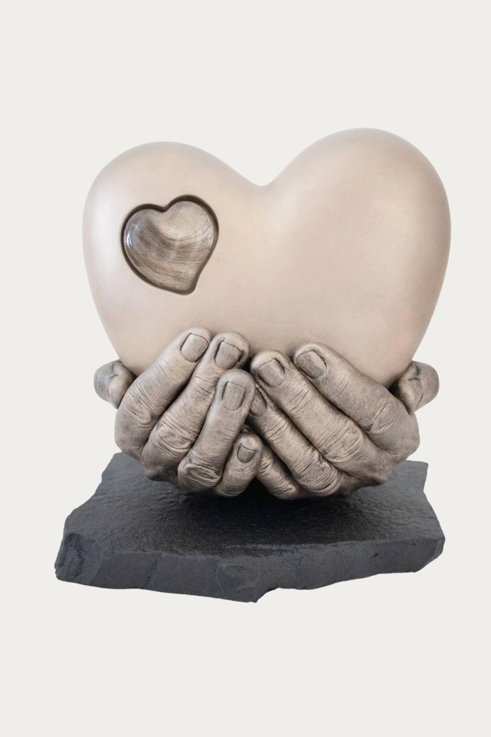 Handgedragen urn in hartvorm, gedragen in handen, met een op glas lijkend hartje waarin een plukje haar is verwerkt.